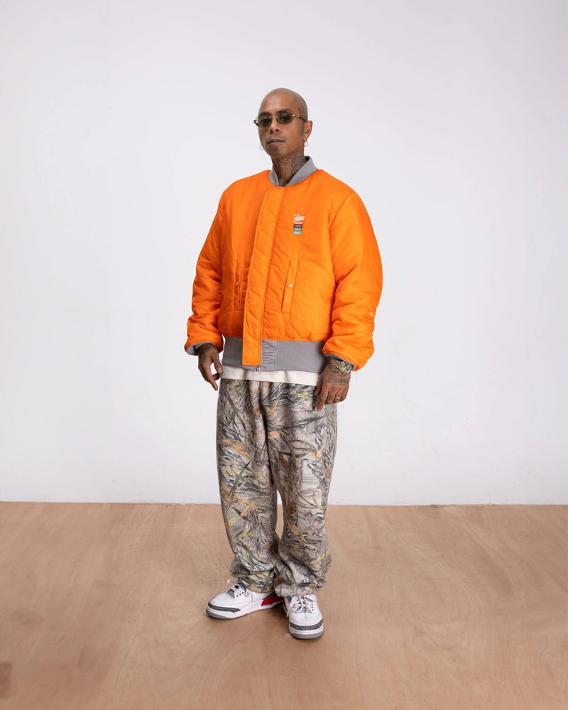 Model wears the Patta x Alpha Industries MA-1 Celebration bomber jacket in orange with camo pants.