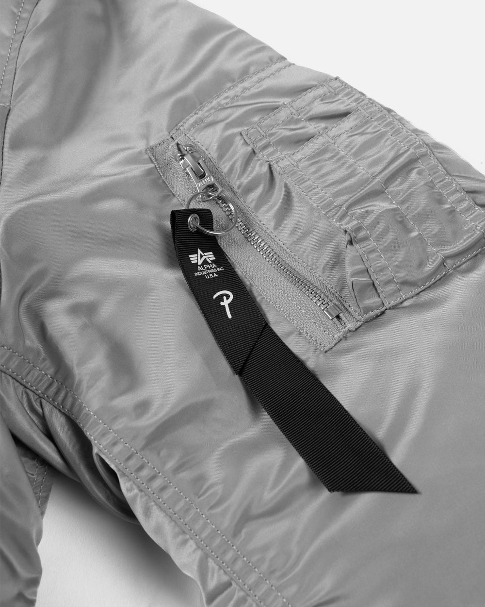 Sleeve pocket detail on the silver Patta x Alpha Industries MA-1 jacket with black branded ribbon tag.