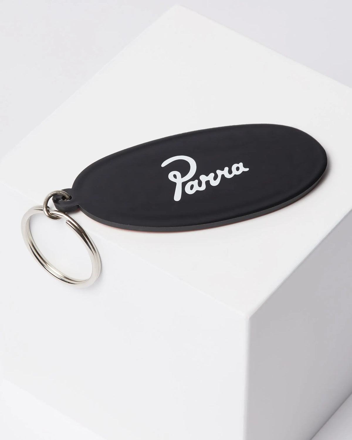 Black oval By Parra rubber keychain with white script logo resting on a white block.