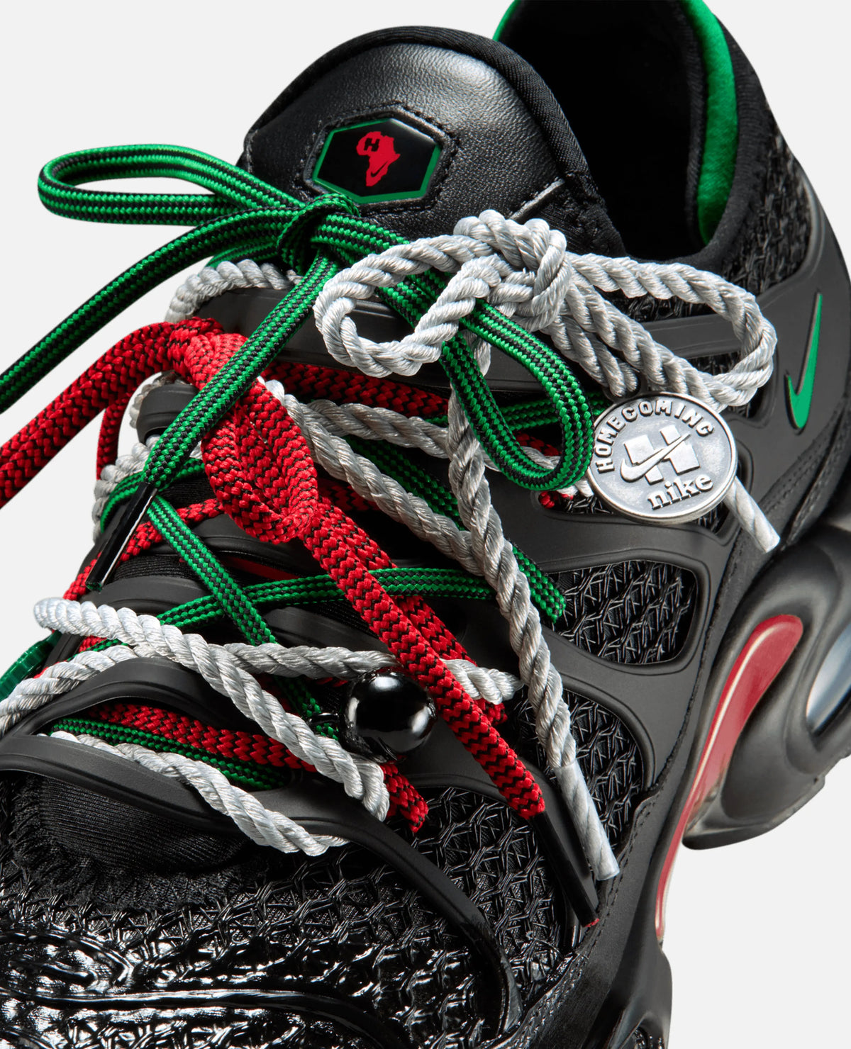 Close-up of HOMECOMING x Nike Air Max Plus laces in red, green, and silver rope with a metallic logo charm.