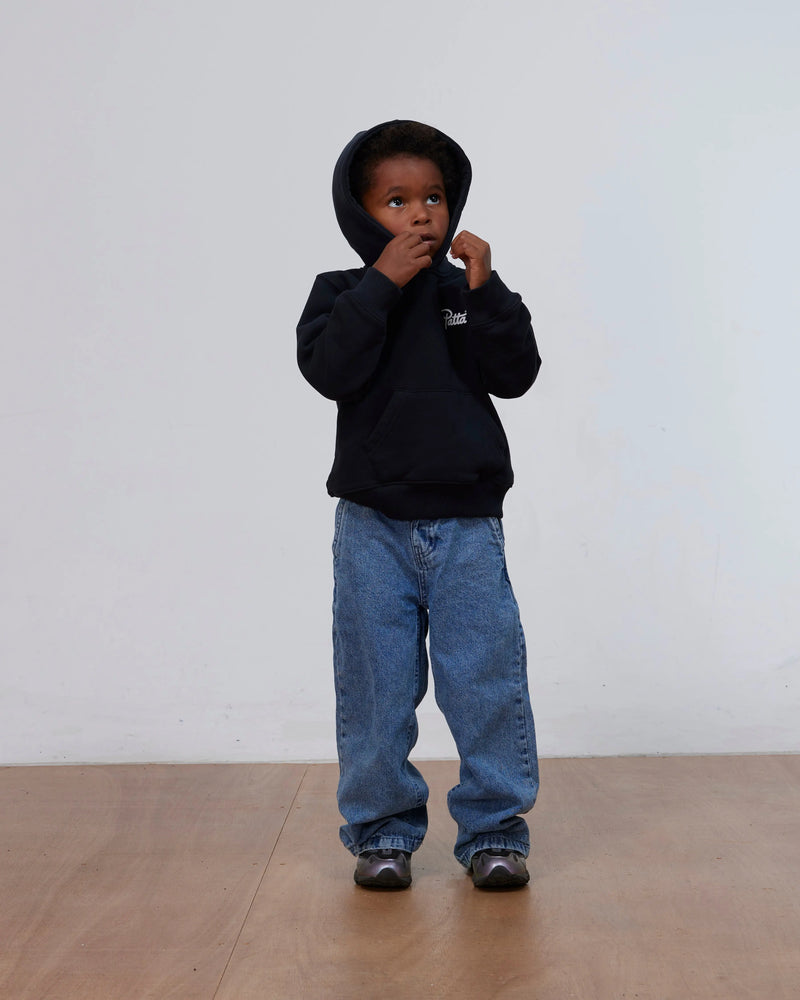 Young model wearing the black Patta Kids Championship Hooded Sweater with blue jeans.