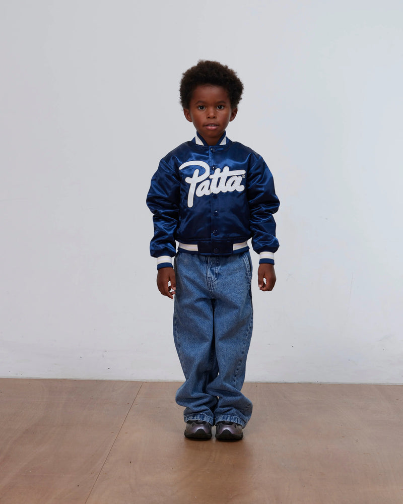 Boy wearing the Patta Kids Satin Bomber Jacket in Black Iris paired with denim jeans in a studio.