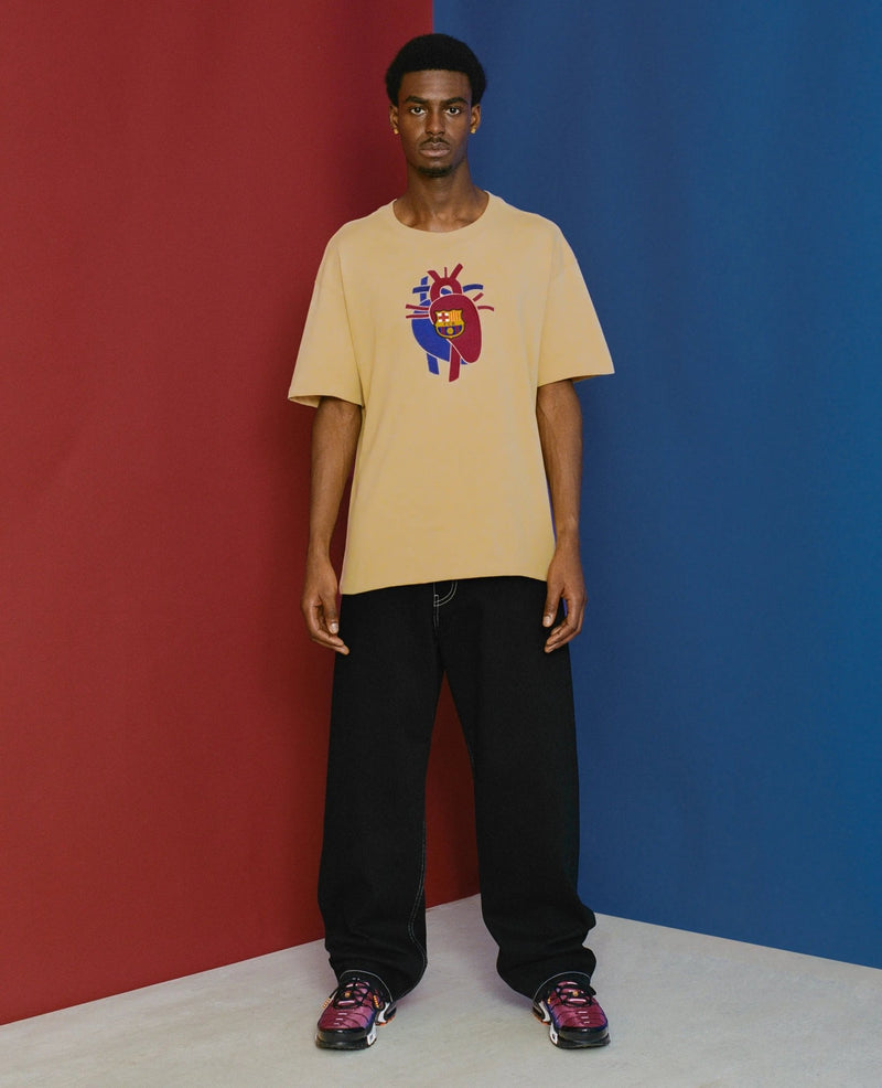 Model wearing the sesame FCB x Patta Culers del Món T-shirt featuring an FC Barcelona heart graphic.