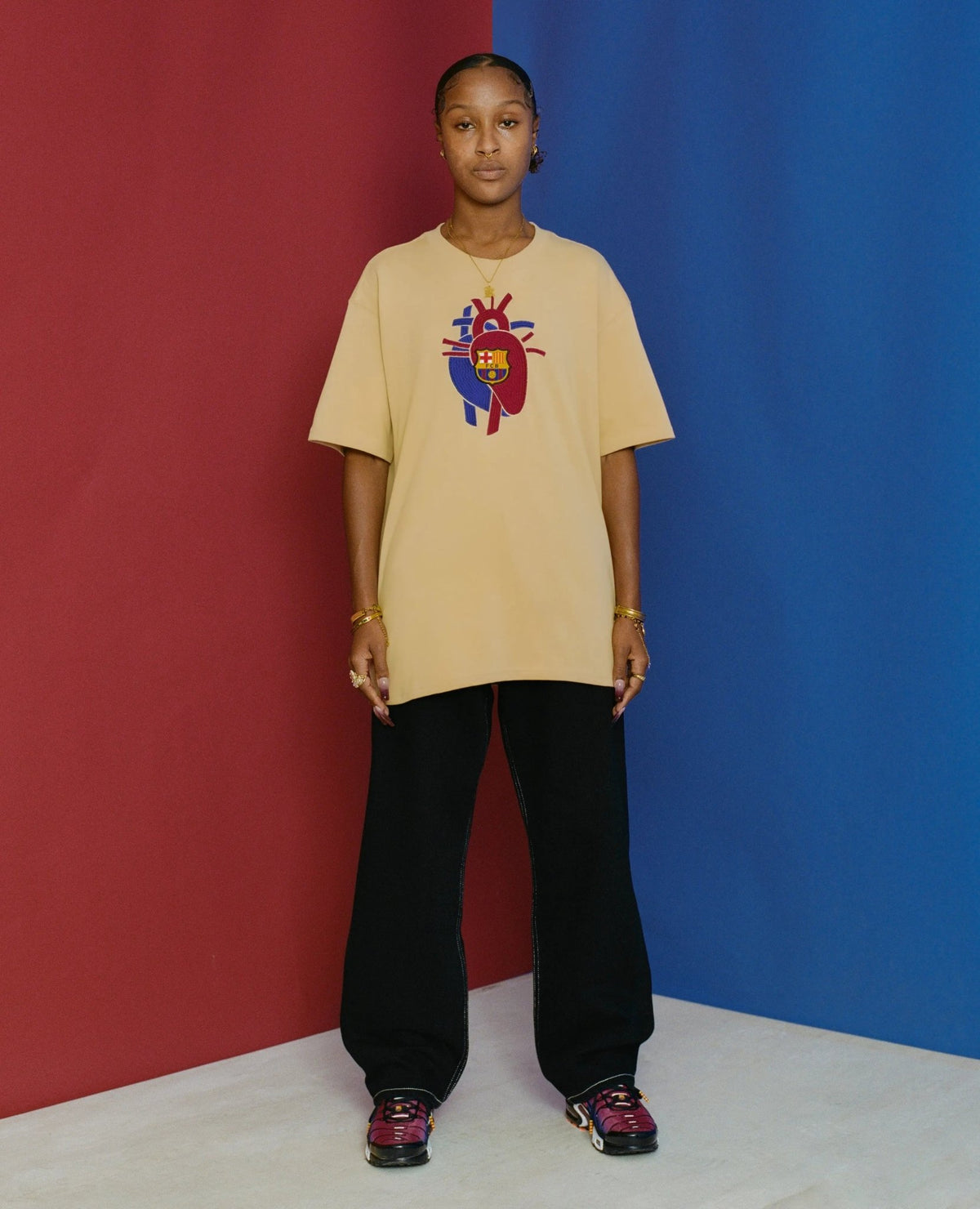 Model wearing the sesame FCB x Patta Culers del Món t-shirt against a split red and blue background.