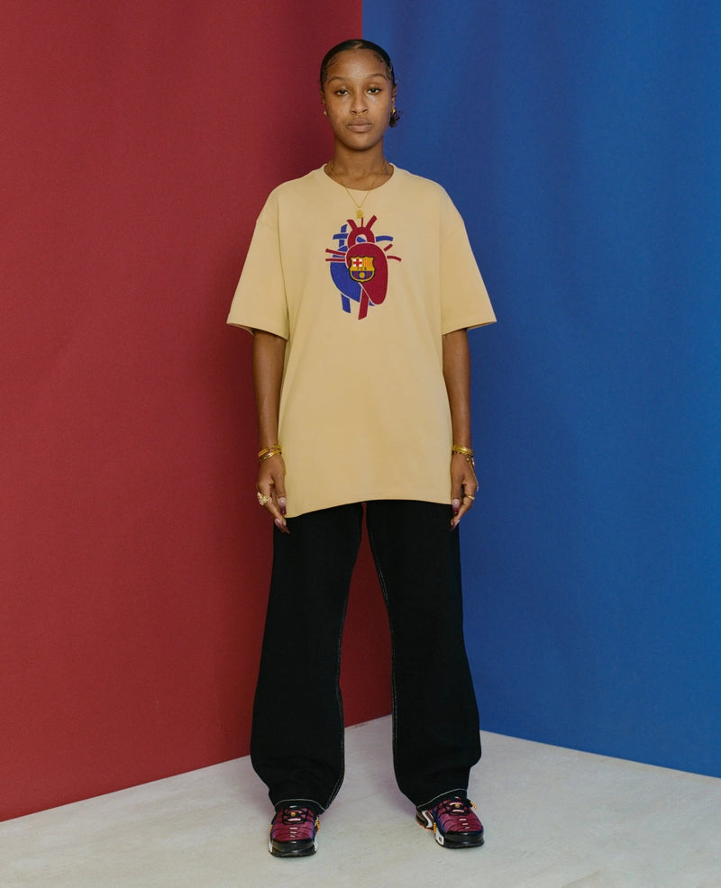 Model wearing the sesame FCB x Patta Culers del Món t-shirt against a split red and blue background.