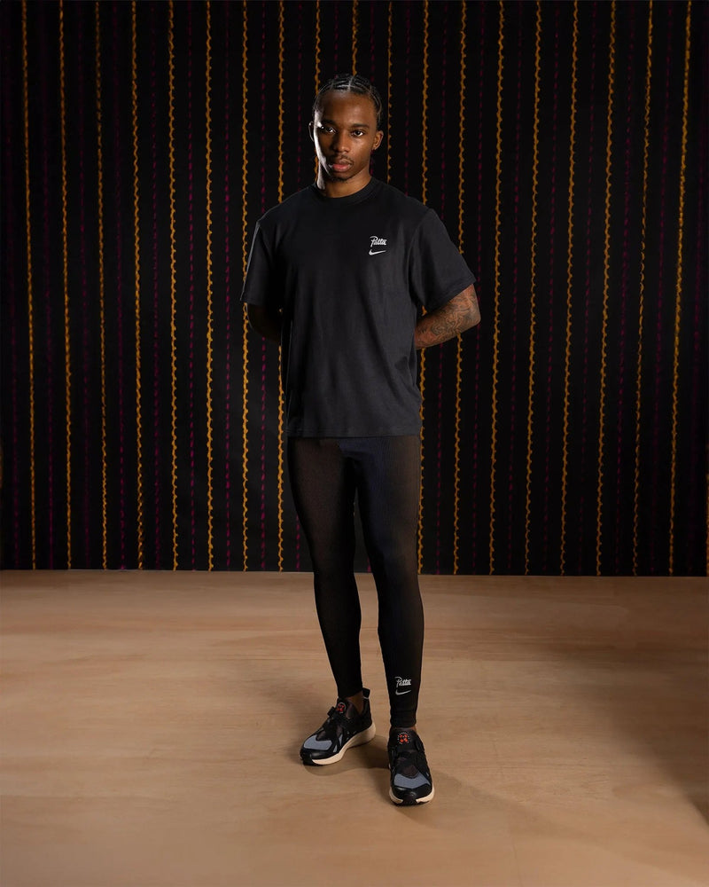 Male model wearing black Nike x Patta Running Team leggings and t-shirt against a dark backdrop.