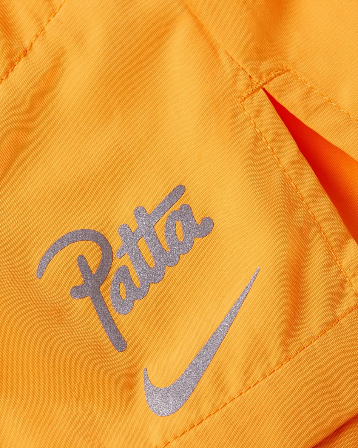 Close-up of reflective Nike Swoosh and Patta logo on orange Nike x Patta Running Team shorts.