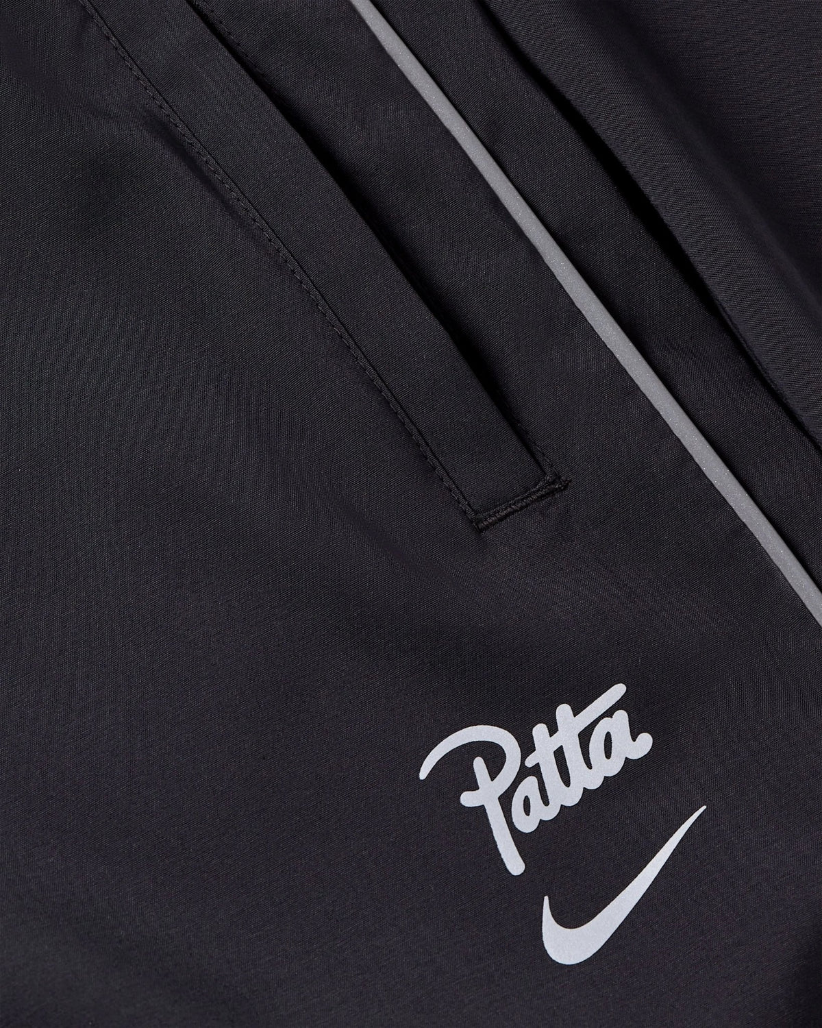 Close-up view of black Nike x Patta Running Team track pants with reflective piping and printed logos.