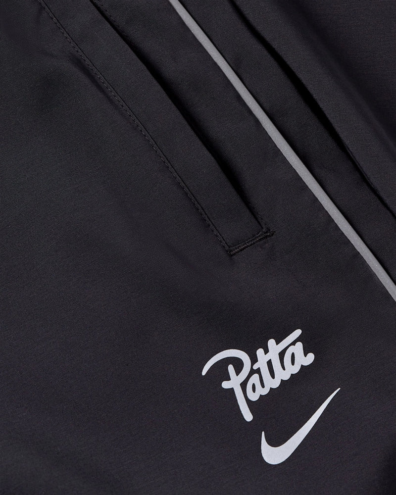 Close-up view of black Nike x Patta Running Team track pants with reflective piping and printed logos.
