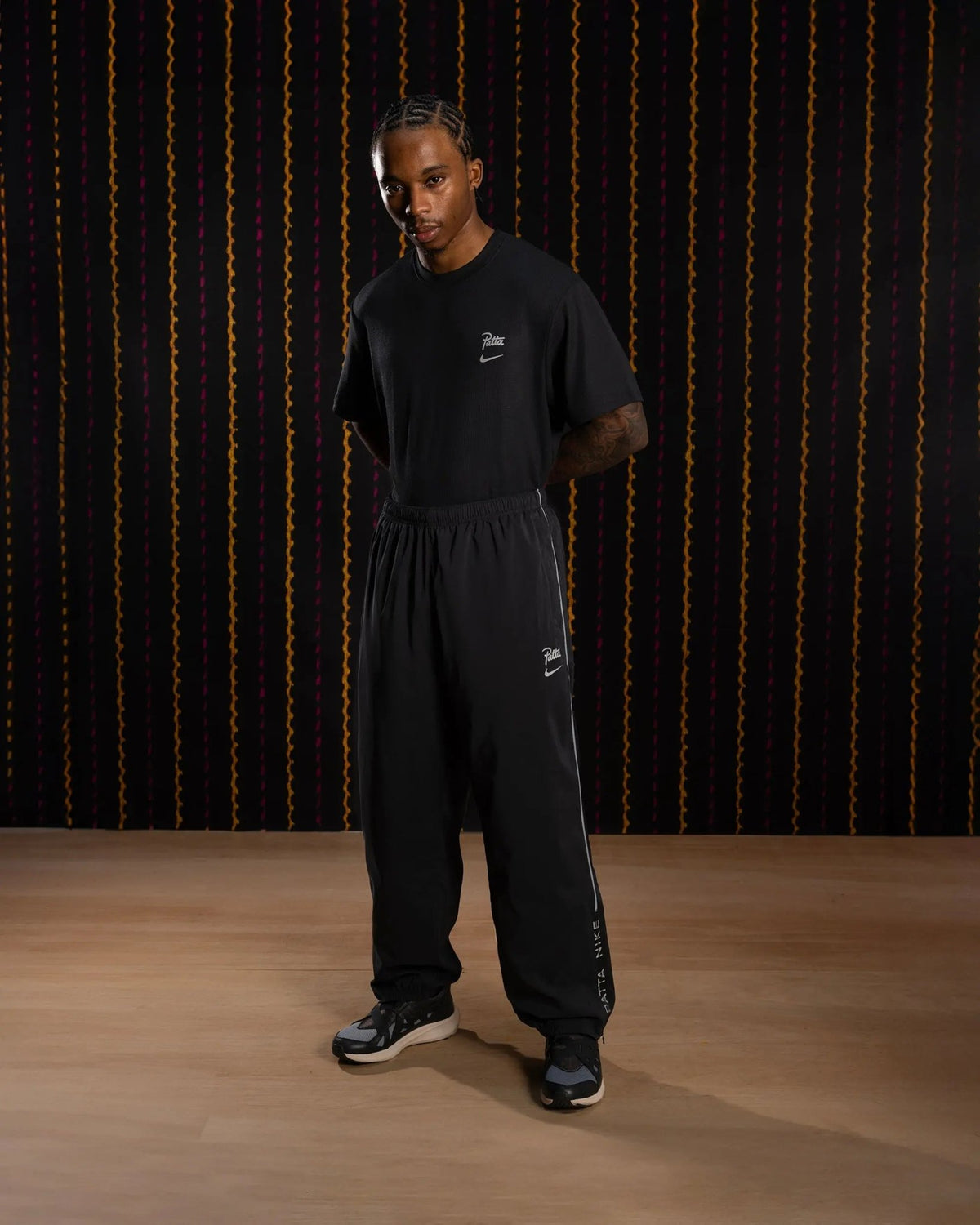 Model wearing black Nike x Patta Running Team track pants with reflective piping and logo details.