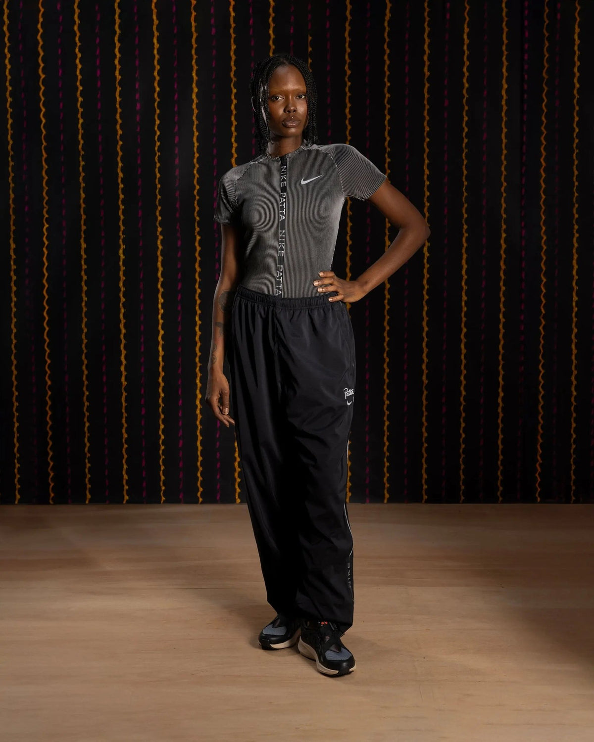 Model wearing black Nike x Patta Running Team track pants against a patterned studio backdrop.