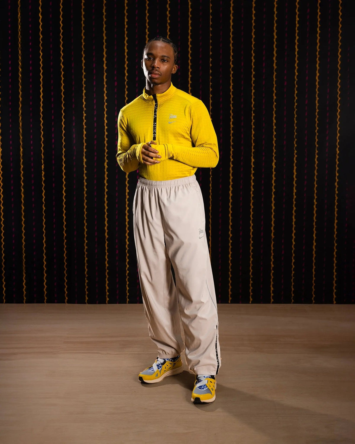 Male model wearing Nike x Patta Running Team track pants in Sanddrift Cream II with a yellow running top.