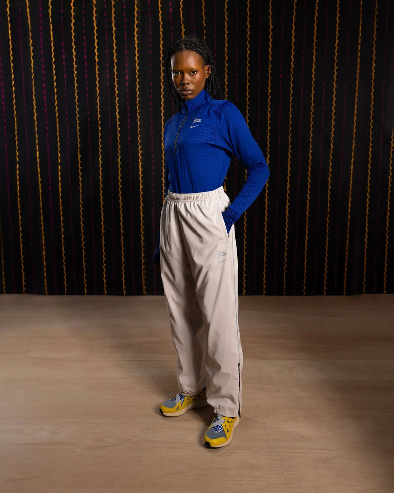 Model wearing Nike x Patta Running Team track pants in Sanddrift and Cream II with a blue half-zip top.