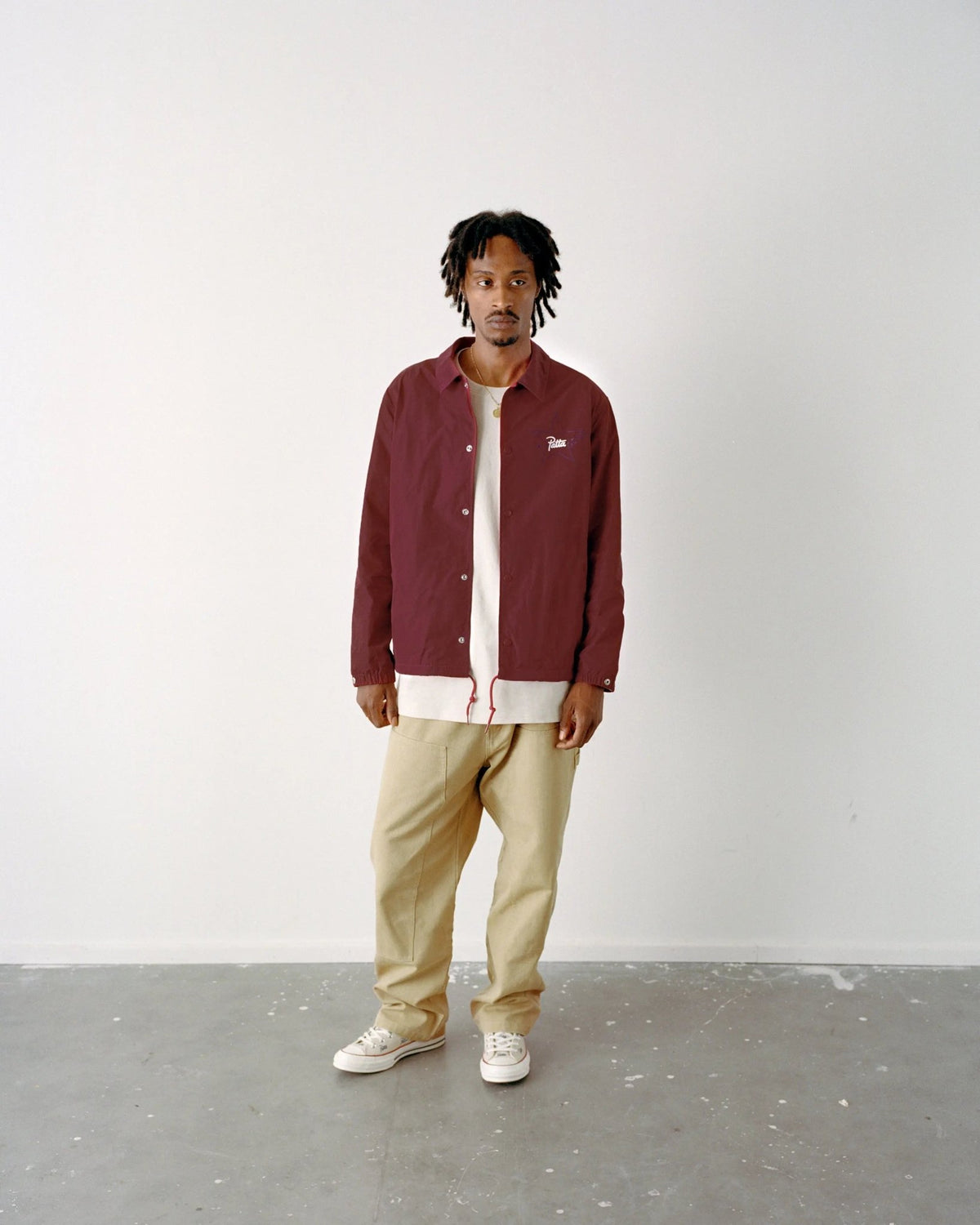 Model wearing the garnet Patta x Converse coaches jacket paired with beige pants in a studio setting.