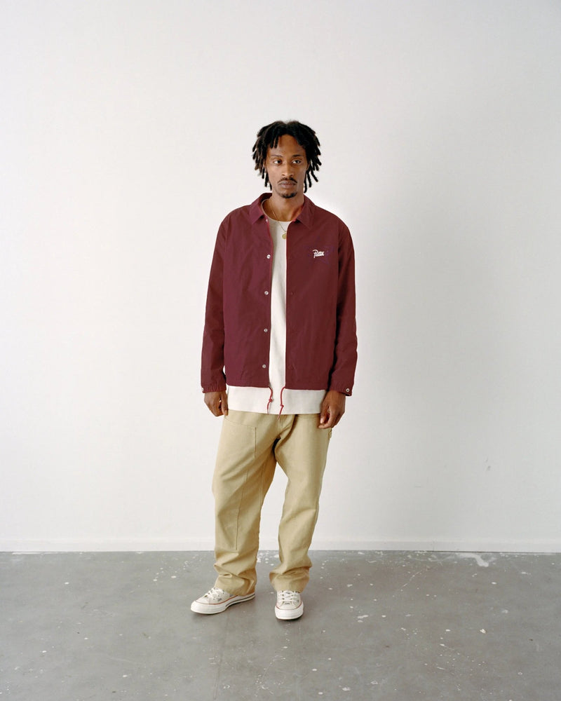 Model wearing the garnet Patta x Converse coaches jacket paired with beige pants in a studio setting.