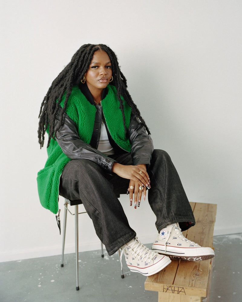 Model wearing the Patta x Converse Jolly Green reversible vest layered over a black jacket.