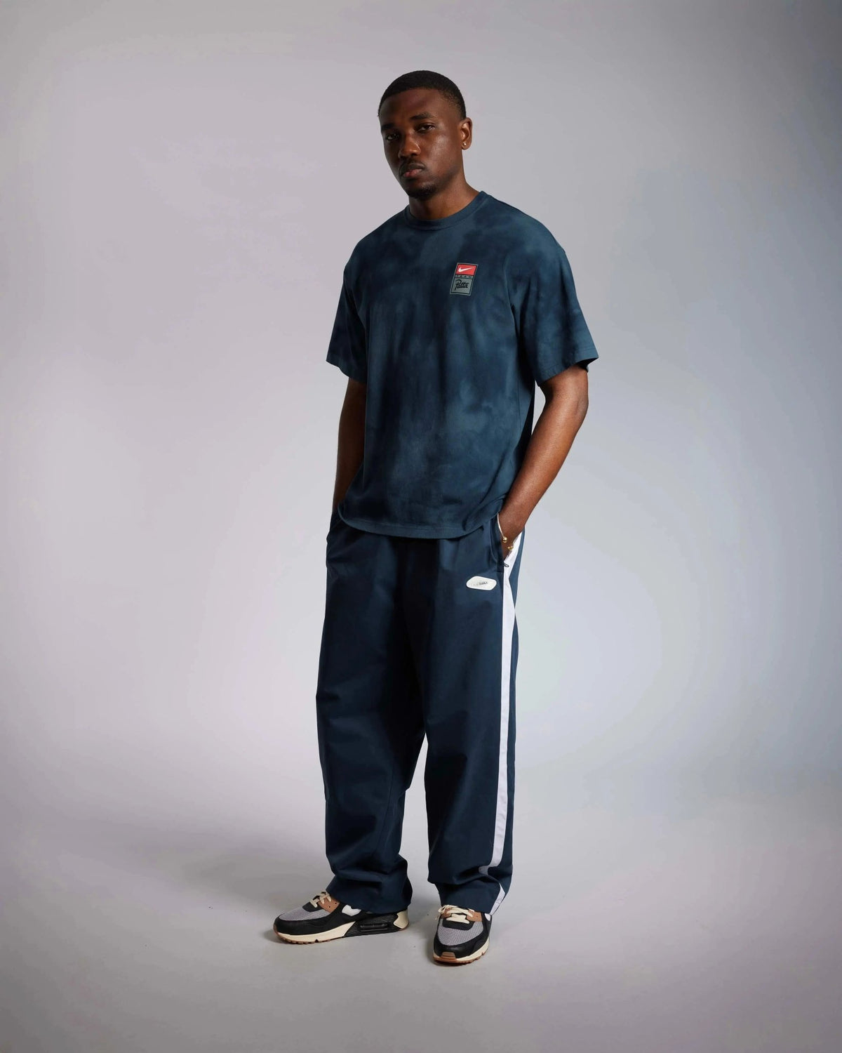 Male model wearing the washed dark blue Patta x Nike NRG T-shirt with navy track pants in a studio shot.